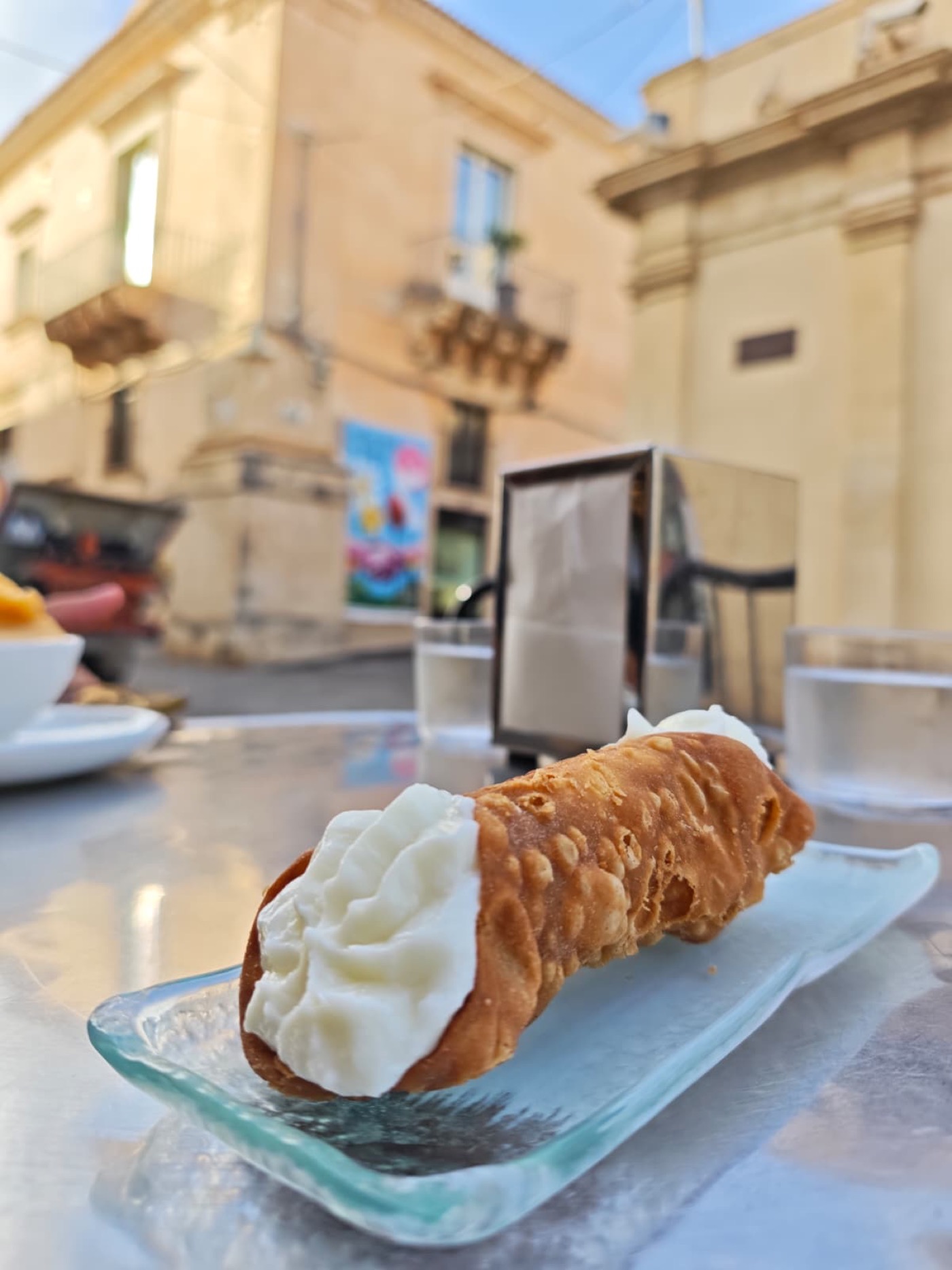 A fresh cannolo from Caffe Sicilia on a glass plate, with Noto's Baroque facades in the background